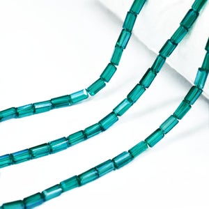 May include: Close-up of strands of teal rectangular glass beads. The beads are strung together, creating a flowing, linear pattern against a white background. The beads have a slightly iridescent quality, reflecting light.