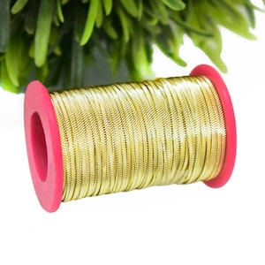 Pode incluir: Uma bobina de cordão metálico dourado enrolada em torno de uma bobina de plástico rosa. O cordão é brilhante e tem uma textura sutil.