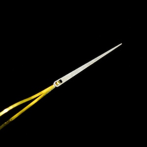 Puede incluir: Primer plano de una aguja plateada con punta afilada y un pequeño ojo. Dos hebras de hilo amarillo están enhebradas en el ojo de la aguja. El fondo es negro.