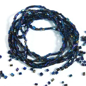 Puede incluir: Una hebra de cuentas de vidrio azul y morado iridiscente. Las cuentas son pequeñas y cilíndricas, y están dispuestas en una espiral suelta. Algunas cuentas están dispersas alrededor de la espiral.