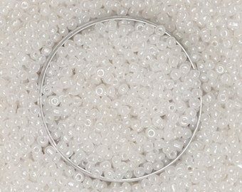 May include: A close-up image of a white opaque seed bead, with a silver metal container in the center. The beads are small and round, and they are arranged in a random pattern.