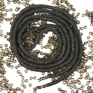 Puede incluir: Primer plano de cuentas de semillas de color bronce oscuro enrolladas en espiral sobre fondo blanco. Cuentas sueltas dispersas. Ideal para la creación de joyas y manualidades.