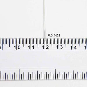 May include: A close-up of a ruler with millimeter markings and a thin, silver wire. The text "0.5 MM" is visible. The wire is positioned at the 12 mm mark on the ruler, indicating its measurement.