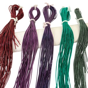 Puede incluir: Cinco carretes de hilo de colores, incluyendo rojo, morado, verde y verde oscuro, cuelgan de una superficie blanca. El hilo está dispuesto en línea vertical, con el hilo rojo a la izquierda y el hilo verde oscuro a la derecha.