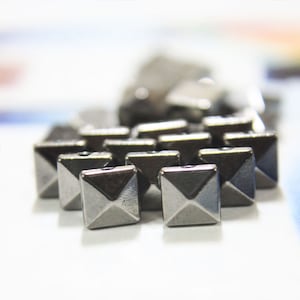 May include: A collection of small, square, silver-coloured metal studs with a pyramid design. The studs are clustered together, some in focus, others blurred. Each stud has a hole for attaching to fabric. The studs are metallic.