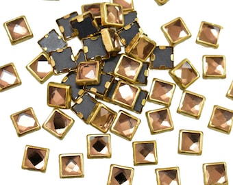 May include: A pile of gold-rimmed, square-shaped, clear glass rhinestones with a light brown tint. The rhinestones are scattered on a white background.