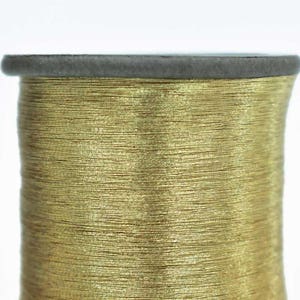 May include: A spool of gold metallic thread. The thread is wound tightly around the spool and has a shiny, metallic finish.