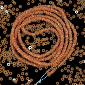 Pode incluir: Um fio de contas redondas cor de coral, cada conta é um disco plano com um furo no centro. As contas estão dispostas em um padrão circular, criando um efeito de camadas. As contas estão espalhadas sobre um fundo preto.