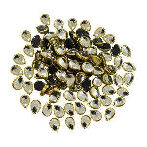 May include: A large pile of clear teardrop-shaped glass rhinestones with gold settings. The rhinestones are arranged in a random pattern.