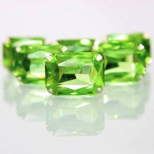 Op de afbeelding: Close-up van verschillende heldergroene, rechthoekige edelstenen met zilveren tanden. De edelstenen zijn facetgeslepen, reflecteren licht en zijn gerangschikt op een reflecterend oppervlak. De edelstenen zijn een levendig, opvallend groen.