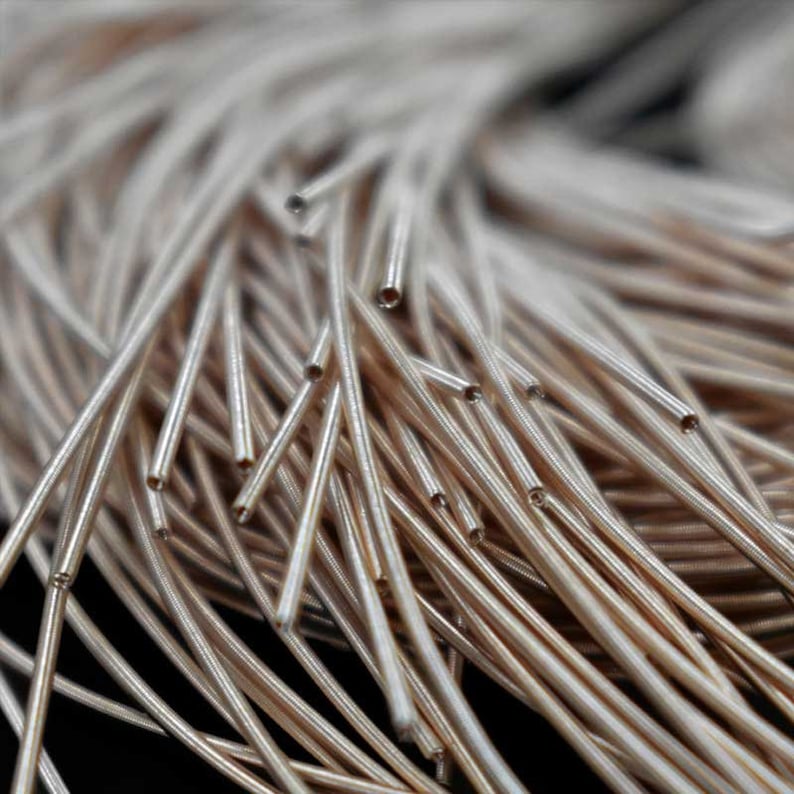 May include: Close-up of a collection of thin, metallic tubes. The tubes are a light bronze color and appear to be made of metal. They are clustered together, with some tubes overlapping others. The ends of the tubes are visible, showing a hollow interior.