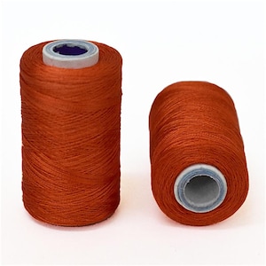 May include: Two spools of orange thread. The thread is wound around a white plastic spool with a grey plastic cap.