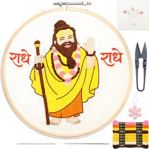 May include: An embroidery kit featuring a wooden hoop with a stitched figure of a smiling bearded person in yellow and orange robes, holding a staff, with pink and white garlands. The text "राधे" is stitched in red on both sides of the figure.