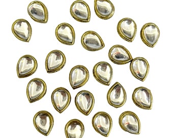 May include: A collection of 25 clear teardrop-shaped glass rhinestones with gold settings. The rhinestones are scattered on a white background.