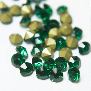 Op de afbeelding: Een verzameling gefacetteerde, smaragdgroene en goudkleurige edelstenen. De edelstenen hebben een veelzijdige slijpvorm, die licht reflecteert. De goudkleurige stenen hebben een matte afwerking. De edelstenen zijn verspreid over een wit oppervlak.