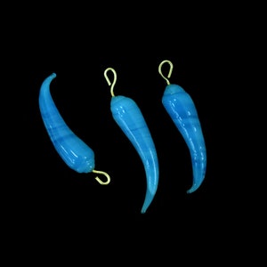 May include: Three blue glass chili pepper pendants with gold-colored wire loops for hanging. The peppers have a curved shape and a translucent appearance, with subtle blue striations. The background is solid black.