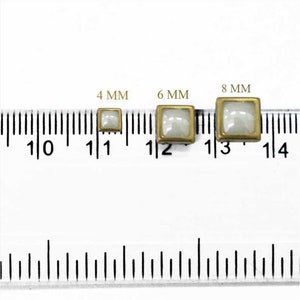 Op de afbeelding: Drie vierkante, parelmoerachtige knopen in wit met goudkleurige metalen frames. De knopen zijn 4 mm, 6 mm en 8 mm groot, zoals aangegeven door de liniaal in de afbeelding.