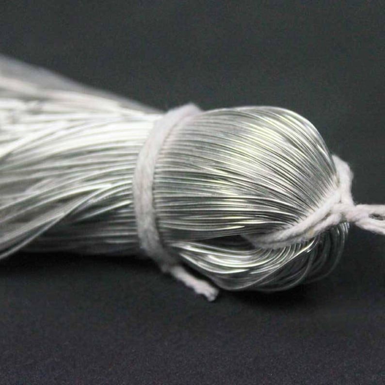 May include: A tightly wound bundle of silver-colored wire, secured with a white string. The wire is neatly coiled, and the string is wrapped around the center, creating a compact package. The background is a dark, solid color.