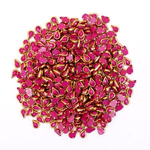 Puede incluir: Un montón grande de piedras de imitación de color rosa en forma de lágrima con engastes dorados. Las piedras de imitación están esparcidas sobre un fondo blanco.