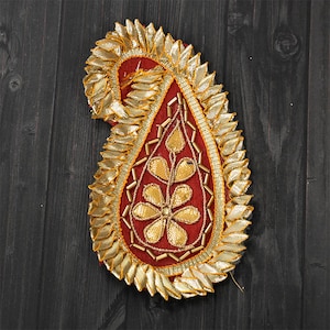 May include: A decorative paisley-shaped applique with a red center and gold embellishments. The applique features a gold leaf-like border and gold floral and beaded accents. The design is set against a dark wood grain background.