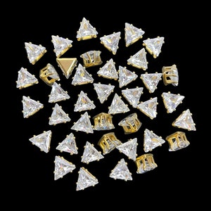 May include: A selection of gold-coloured metal nail art studs, each featuring a clear, triangular-cut rhinestone. The studs are arranged on a black backdrop, highlighting their sparkling facets and detailed design. Some studs have a cage-like structure.