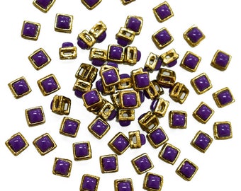 Op de afbeelding: Een strooiing van vierkante knopen in paars met gouden metalen frames. De knopen zijn in een willekeurig patroon op een witte achtergrond gerangschikt.