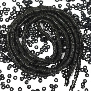 Puede incluir: Una hebra de cuentas redondas, planas y negras. Las cuentas están dispuestas en forma de espiral y están esparcidas sobre un fondo blanco.