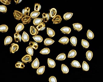May include: A scattering of gold-toned teardrop-shaped embellishments with white pearl centers. The embellishments are scattered on a black background.