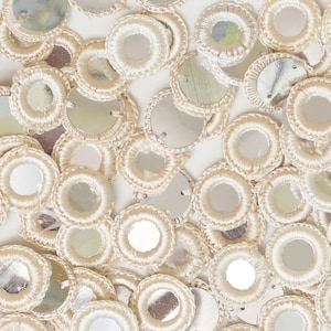 Puede incluir: Primer plano de lentejuelas circulares de color marfil con un centro plateado reflectante. Cada lentejuela está rodeada por un borde bordado de color crema. Las lentejuelas están dispersas por la superficie, creando un elemento decorativo texturizado.