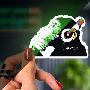 May include: A black and white sticker of a gorilla wearing colorful headphones. The gorilla is in a listening pose with its head tilted to the side. The sticker is on a hand.