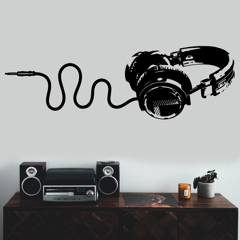 May include: Black and white silhouette of headphones with a cord on a white wall. There are two black speakers and a stereo system on a wooden shelf below the headphones.