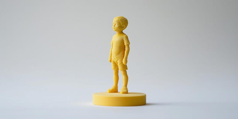 Op de afbeelding: Een gele beeldje van een jong kind, staand op een gele cirkelvormige basis. Het kind draagt een shirt met korte mouwen en een korte broek. Het beeldje is effen van kleur en heeft een gladde textuur. De achtergrond is effen wit.