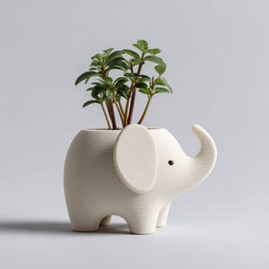 Op de afbeelding: Een matte, gebroken witte keramische plantenbak in de vorm van een gestileerde olifant met een textuur op het oppervlak. De olifant heeft een rond lichaam, vier korte poten, een groot oor en een omhoogstaande slurf. Een kleine groene plant groeit bovenin.