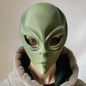May include: A green alien mask with large eye openings and a smooth, textured surface. The mask has a distinctive forehead shape and a neutral expression. The person wearing the mask is wearing a gray hooded jacket.