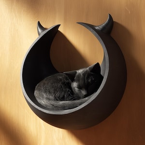 Puede incluir: Un gato negro durmiendo dentro de una cama para gatos en forma de media luna de color gris oscuro. La cama está montada en una pared de madera marrón claro. La cama tiene dos detalles curvos en forma de cuerno en la parte superior.