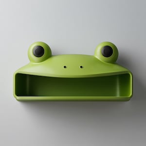 May include: A bright green frog-shaped wall shelf. The shelf features a rectangular storage area, two large, round eyes with black pupils, and two small nostril details. The design is intended for wall mounting.