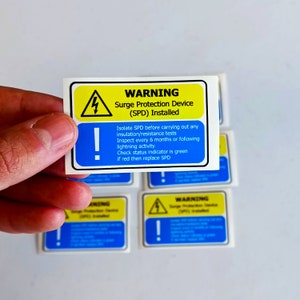 May include: Yellow and blue warning sticker with a lightning bolt symbol and an exclamation point. The text reads "WARNING Surge Protection Device (SPD) Installed Isolate SPD before carrying out any insulation/resistance tests Inspect every 6 months or following lightning activity Check status indicator is green if red then replace SPD"