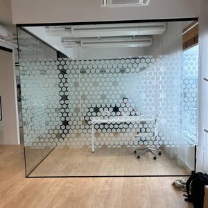 May include: An office space with a glass partition featuring a honeycomb pattern. The room has a white desk, a rolling chair, and wooden flooring. Overhead, there are several air conditioning units.