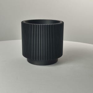 Pu&ograve; includere: Un vaso cilindrico nero opaco caratterizzato da una distinta trama a coste verticali lungo le pareti esterne. Il contenitore presenta un'apertura superiore ampia e una base circolare leggermente rientrata.