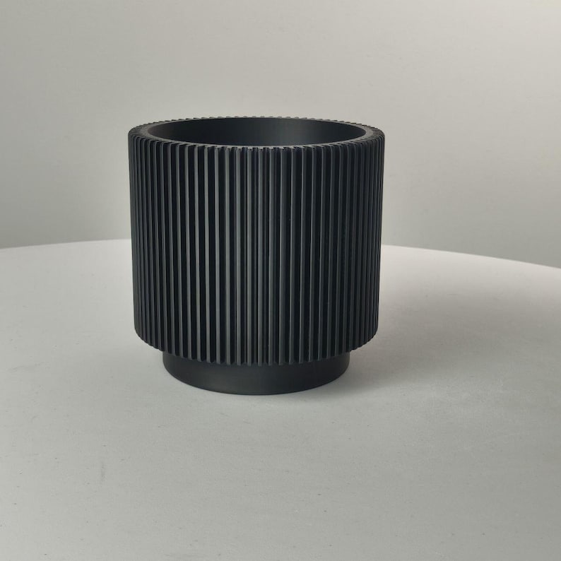 Pu&ograve; includere: Un vaso cilindrico nero opaco caratterizzato da una distinta trama a coste verticali lungo le pareti esterne. Il contenitore presenta un'apertura superiore ampia e una base circolare leggermente rientrata.