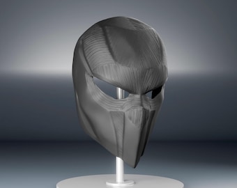May include: A dark gray, futuristic-style helmet with a textured surface and angular design. The helmet features eye openings and a pointed lower section, mounted on a white stand with a circular base. The overall aesthetic suggests a sci-fi or cosplay theme.