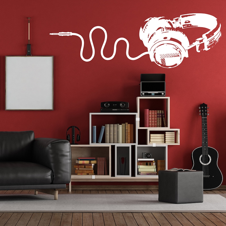 May include: A white silhouette of headphones with a cord on a red wall. The headphones are on a shelf with books and a guitar. There is a black leather couch in the foreground.