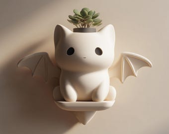 May include: A white cat-shaped planter with bat wings, holding a small succulent plant. The planter has large black eyes and a small, curved mouth. The wings are spread out to the sides. The planter is mounted on a wall.