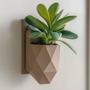 Puede incluir: Una maceta de pared geométrica de color beige con una suculenta artificial. La maceta tiene un patrón de diamantes y está montada sobre un respaldo rectangular. La suculenta tiene hojas grandes y verdes, añadiendo un toque de naturaleza a la decoración.