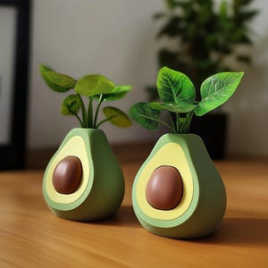 Può includere: Due vasi in ceramica a forma di avocado di colore verde con dettagli gialli e marroni. Ogni vaso contiene una piccola pianta verde.