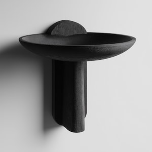 May include: A contemporary, black wooden wall shelf. It has a circular, bowl-shaped top, a curved back, and a sculptural base. The dark colour and minimalist style make it a striking decor piece. Dimensions are approximately 40cm in diameter.