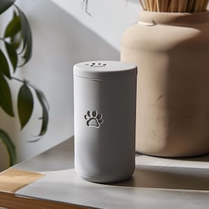 May include: A light grey pet urn with a paw print cutout on the front. The lid also features paw print cutouts. The urn is cylindrical and sits on a light-coloured surface. A beige vase is in the background.