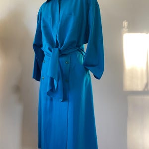 May include: A vibrant turquoise dress with long sleeves and a collared neckline. The dress features a tie at the waist, a button-down front, and a knee-length skirt. The dress is displayed on a mannequin.
