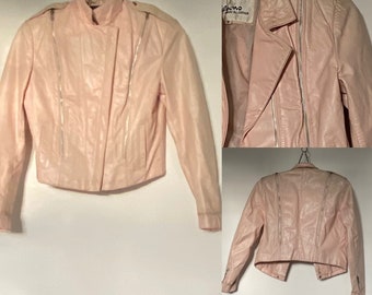 Puede incluir: Una chaqueta de cuero rosa con cierre de cremallera y detalles plateados. La chaqueta tiene un cuello alto y una longitud recortada.