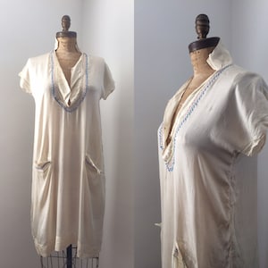 May include: A white cotton tunic dress with short sleeves and a V-neckline. The neckline is decorated with colorful embroidery. The dress has two pockets on the front.
